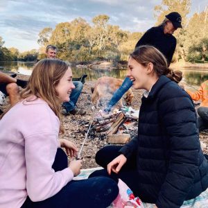2 teenage girld roasting marshmellows by a fire with their parents in the background